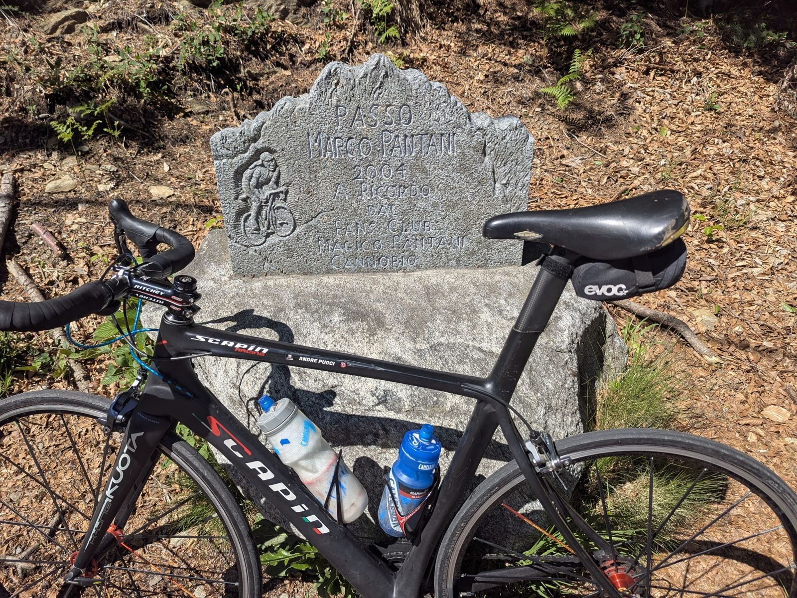 A black road bike leaning against a carved stone memorial reading Passo Marco Pantani at the top of the pass