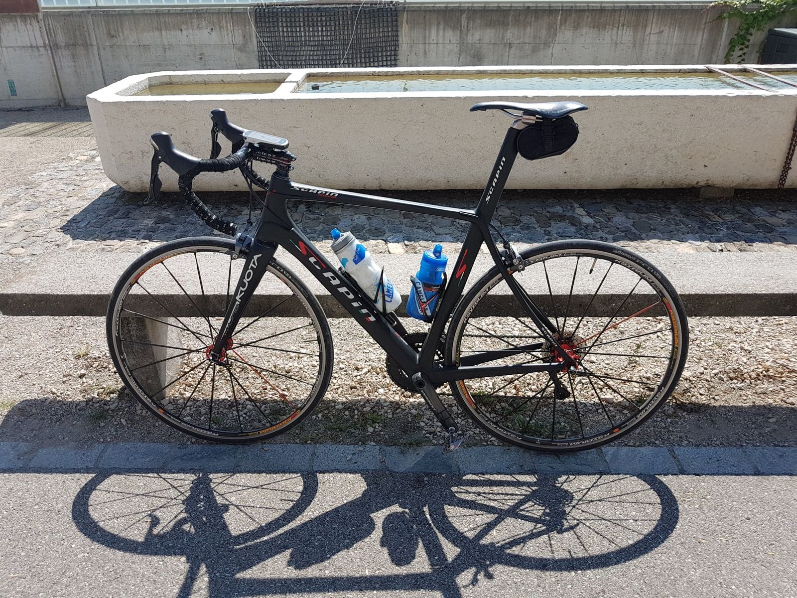 A carbon road bike leaning against a long stone water trough beside the lakeside road at Tüscherz