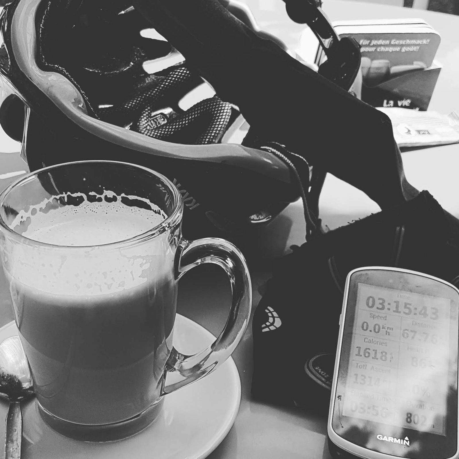 A glass mug of milky coffee next to a Garmin bike computer and a cycling helmet, in black and white