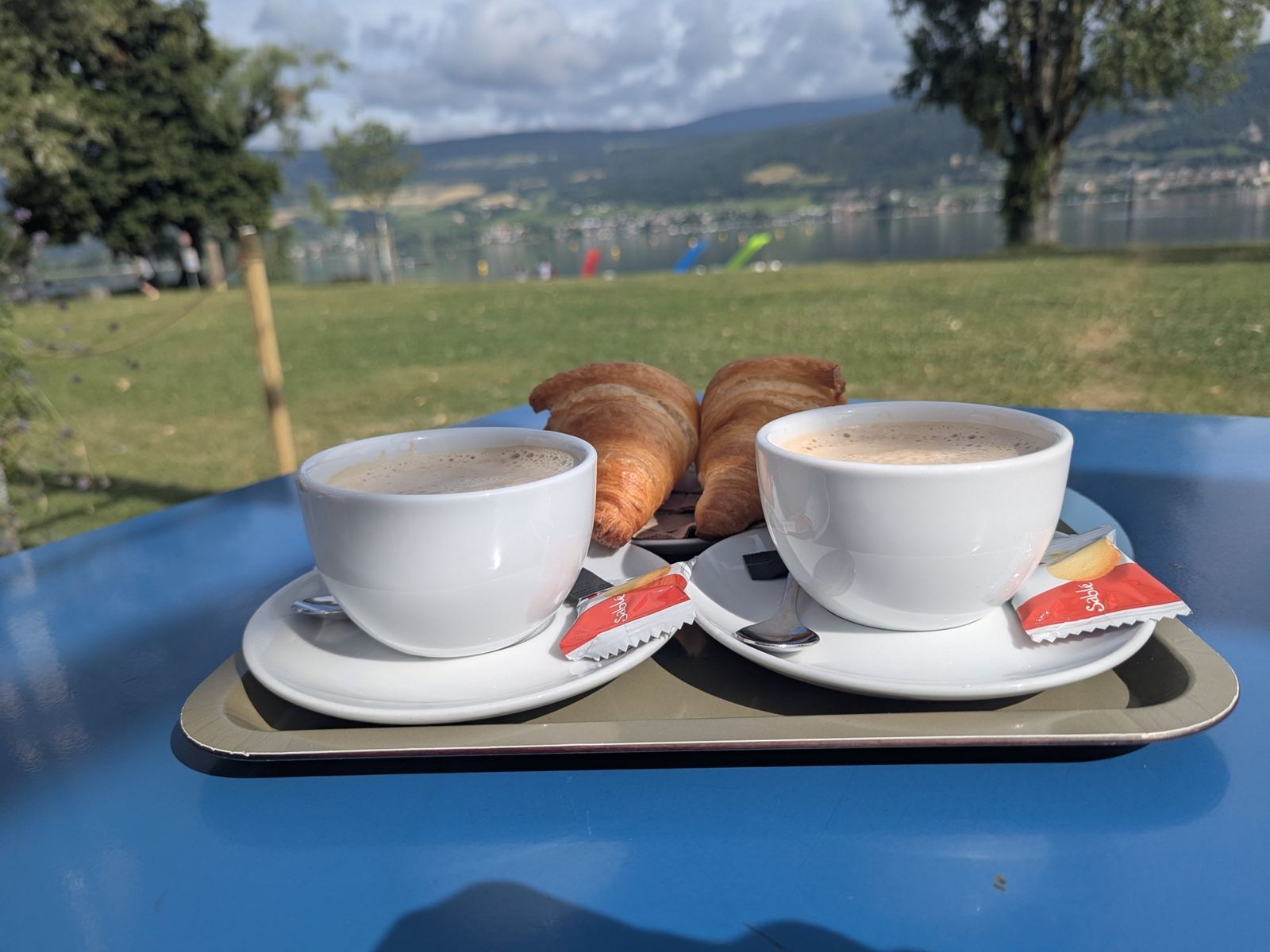 Two cappuccinos and a croissant on a green café table by the water at Erlach