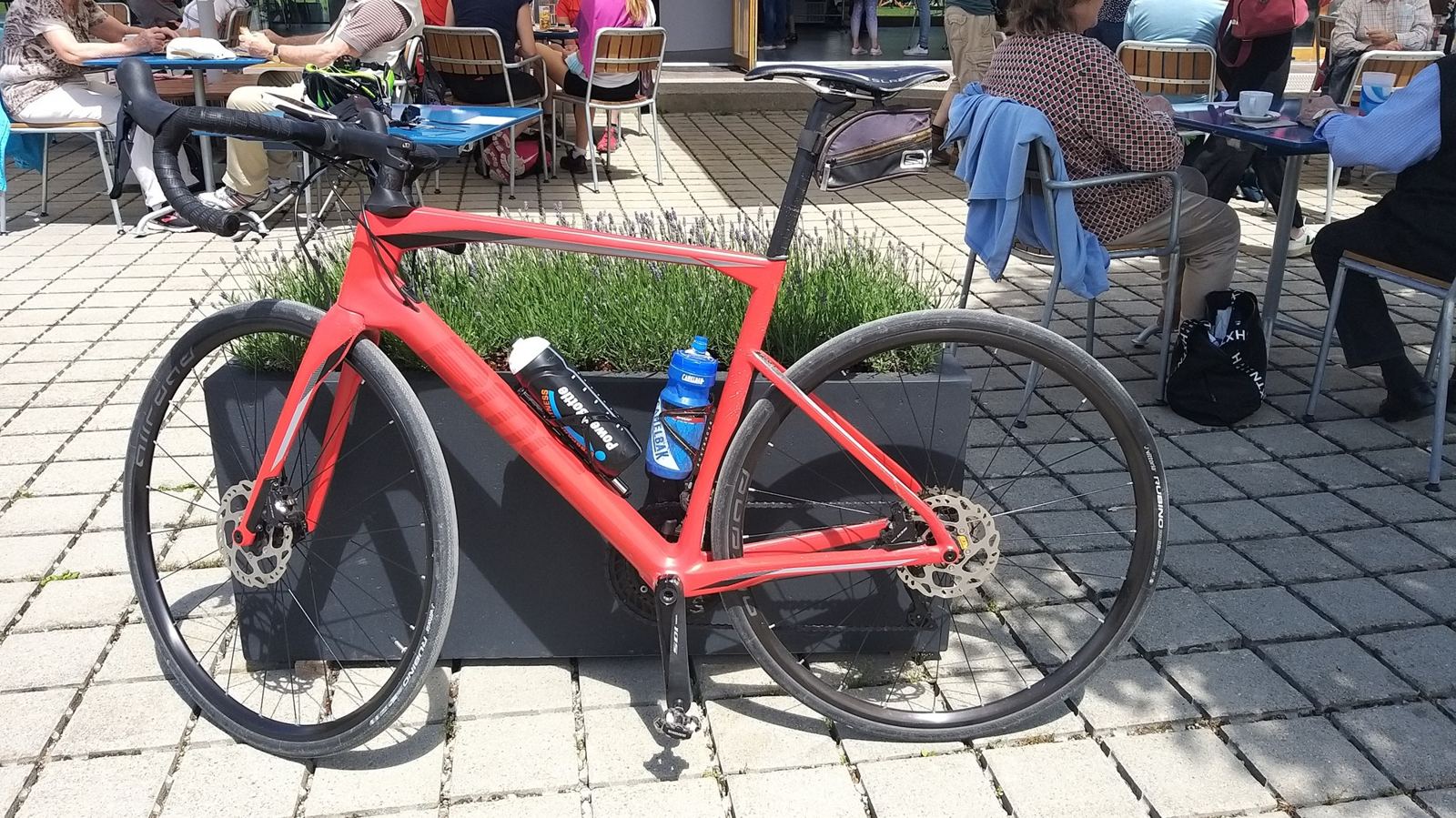 A red carbon gravel bike leaning against a planter outside a busy café terrace in Erlach