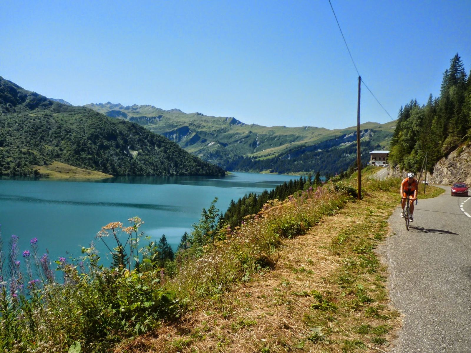The turquoise Lac de Roselend below green mountains, a cyclist on the road above