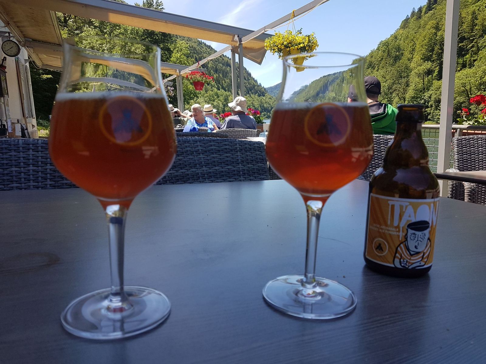 Glasses of beer on a terrace table by the river at La Goule, with diners and wooded hills behind