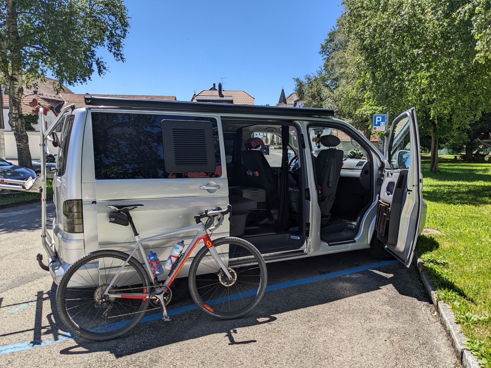A silver VW T5 van parked in Le Noirmont with a red road bike leaning against it