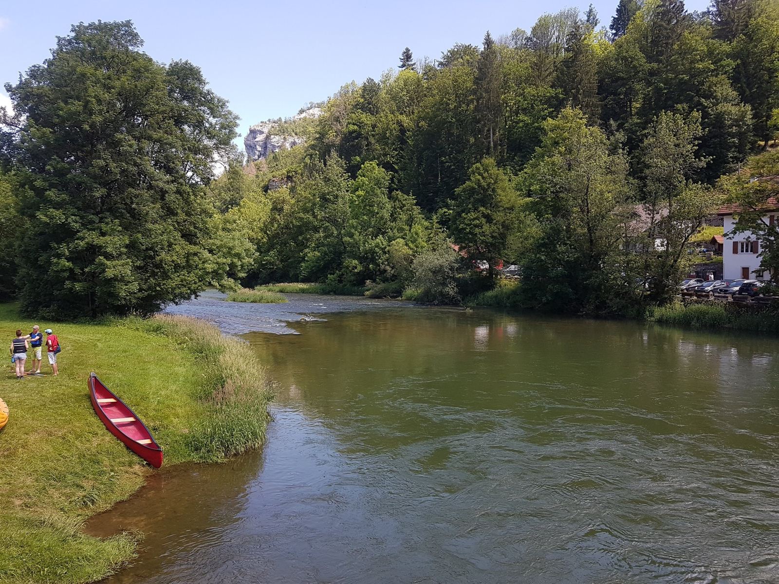 The Doubs at Goumois with a red canoe pulled up on the grass bank and people by the water