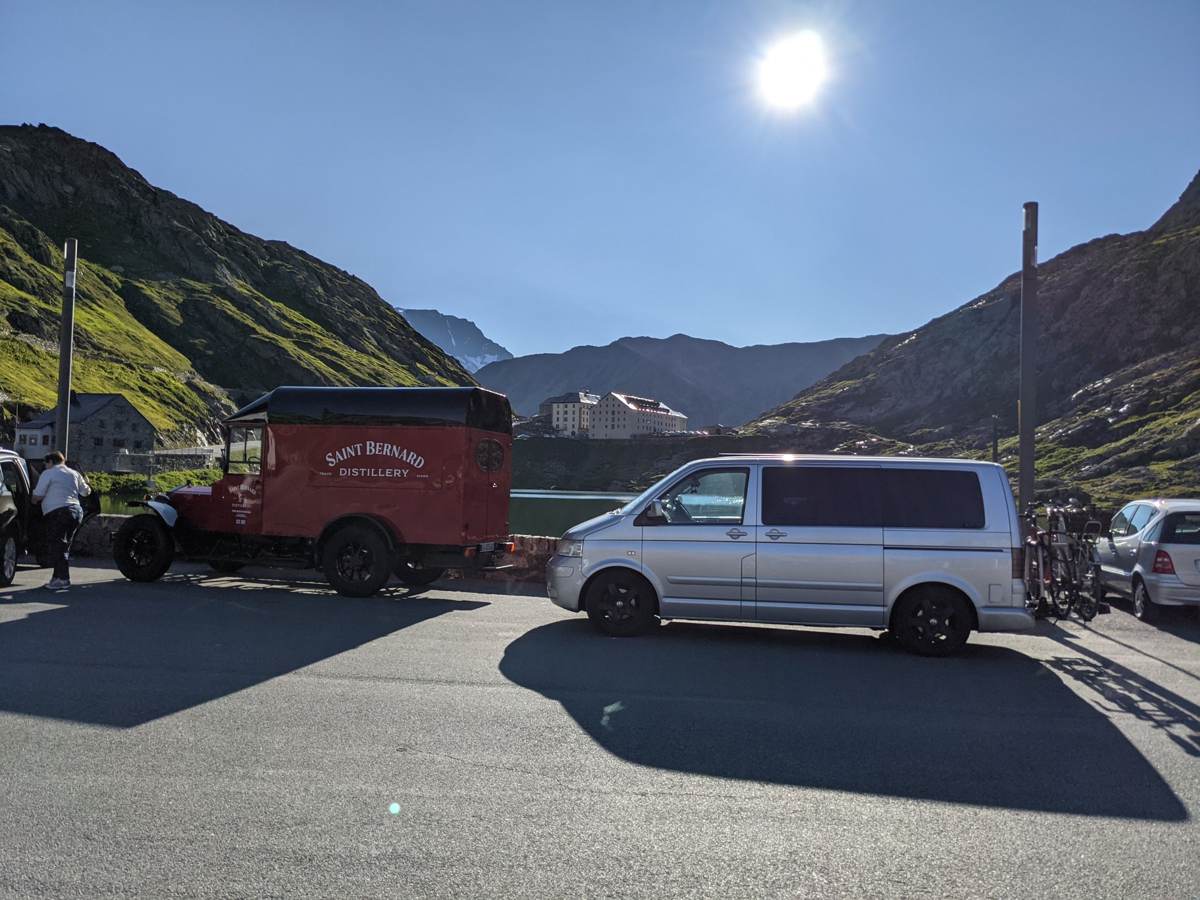 A silver VW T5 van with bikes on the back, parked at the summit of the Grand St Bernard Pass next to a vintage red Saint Bernard Distillery van