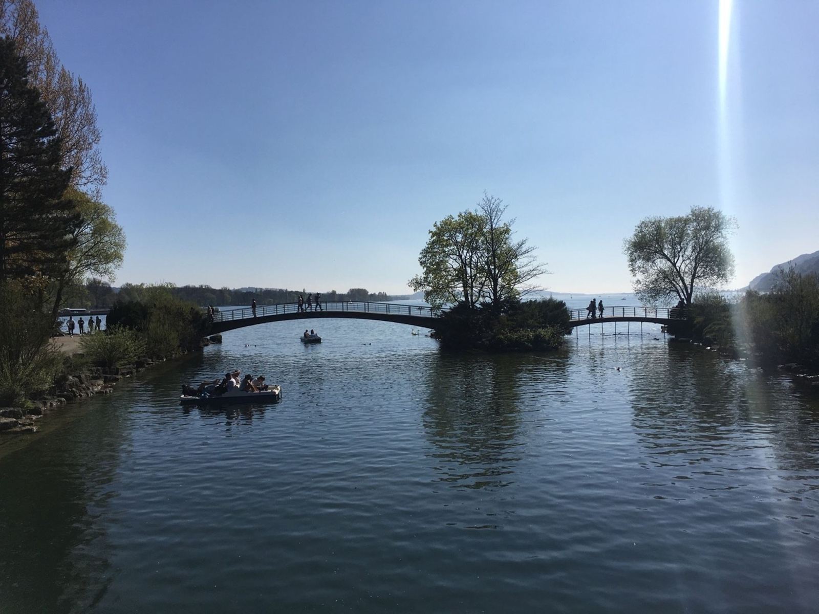 A small footbridge crossing to a little wooded island on Lake Biel, with a boat on the water, in bright April sun