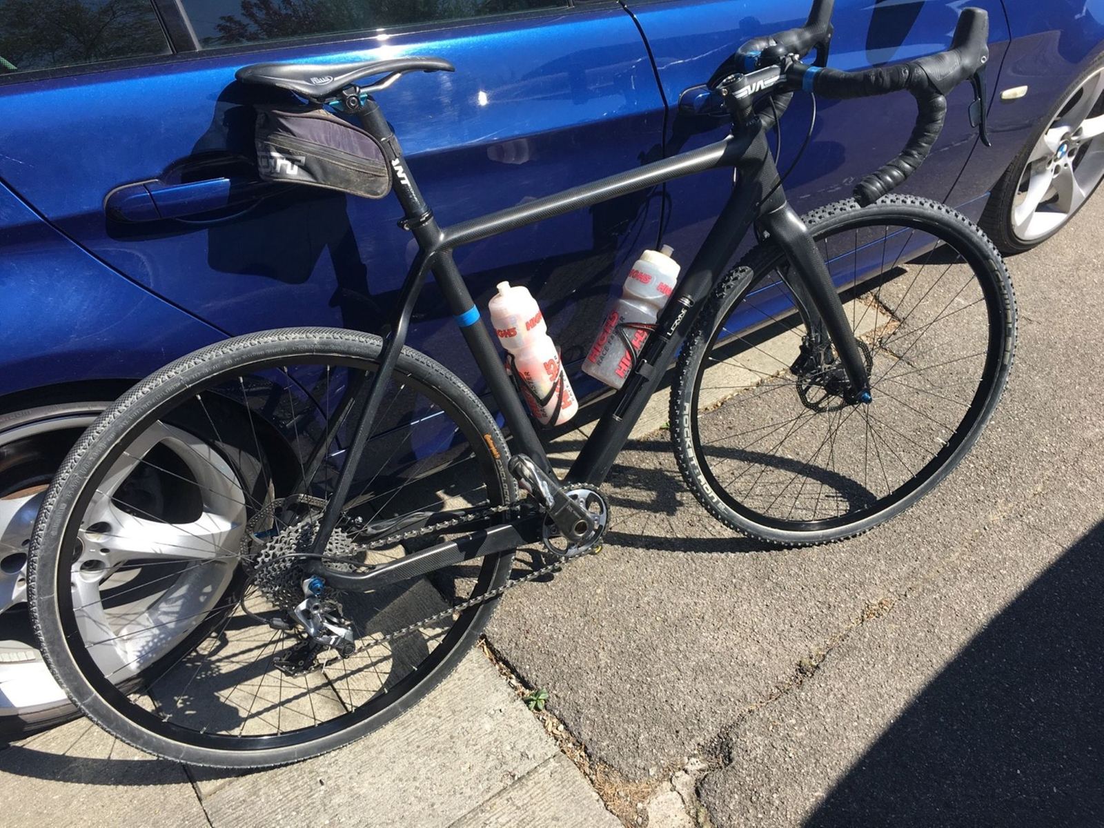 A black carbon cross bike leaning against a blue car on a sunny street