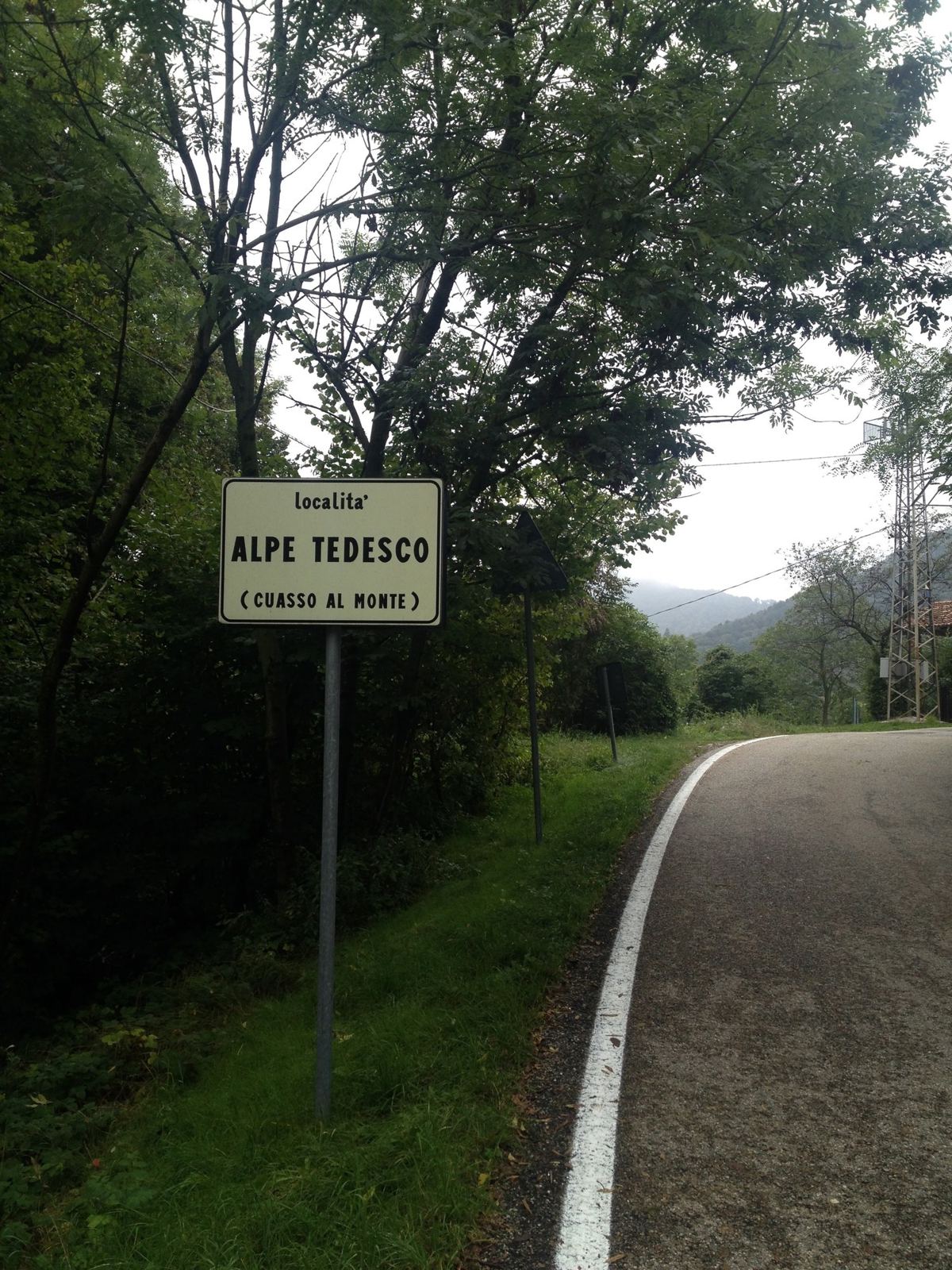 A road sign for Alpe Tedesco beside a quiet road through the woods