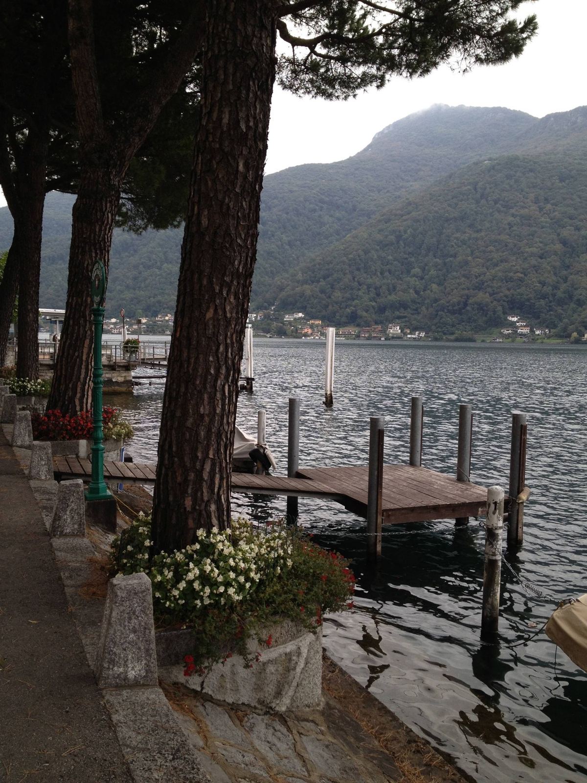 A wooden jetty on Lake Lugano with a lamp post and trees, mountains behind