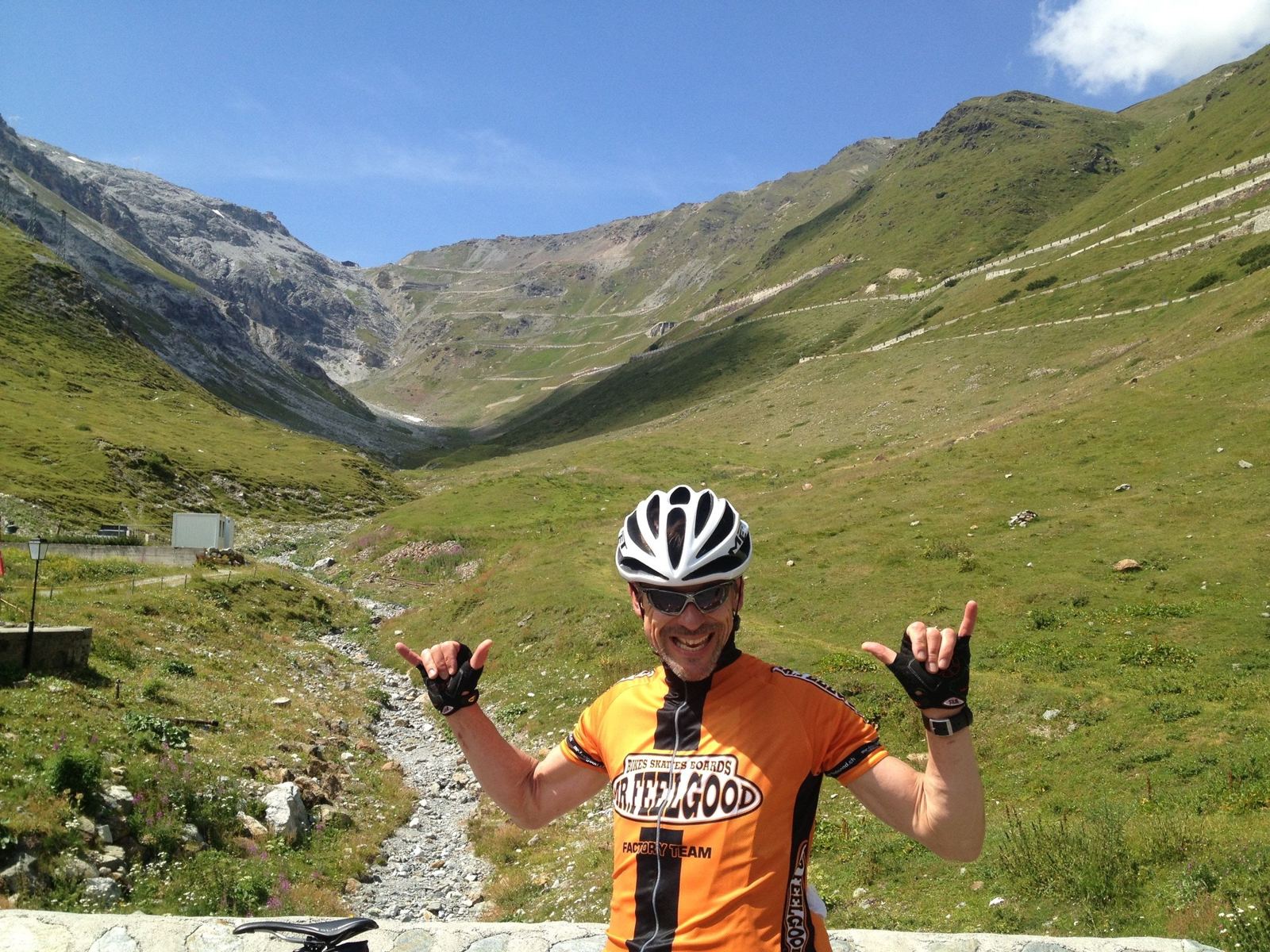 A cyclist in an orange jersey with arms raised on the Stelvio road, with the green valley and mountains behind