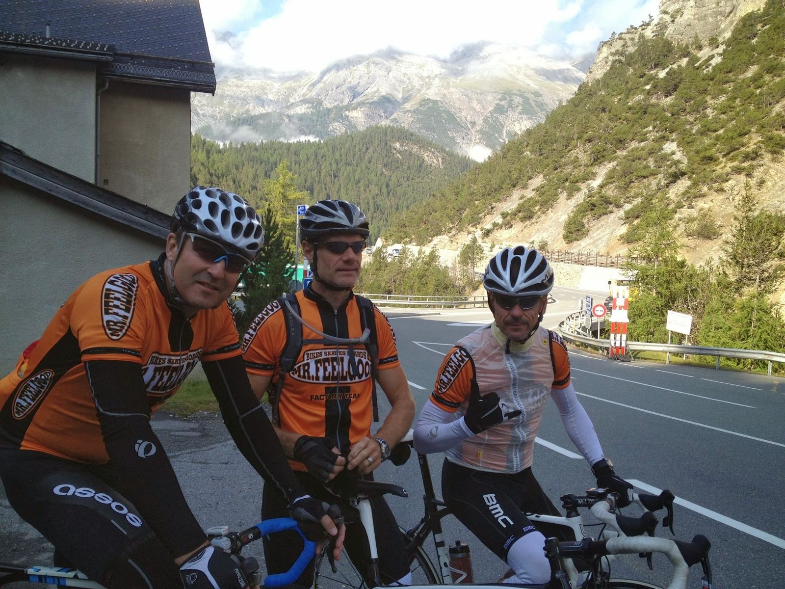 Three cyclists in orange team jerseys at the side of the road at the start of the ride, with mountains behind