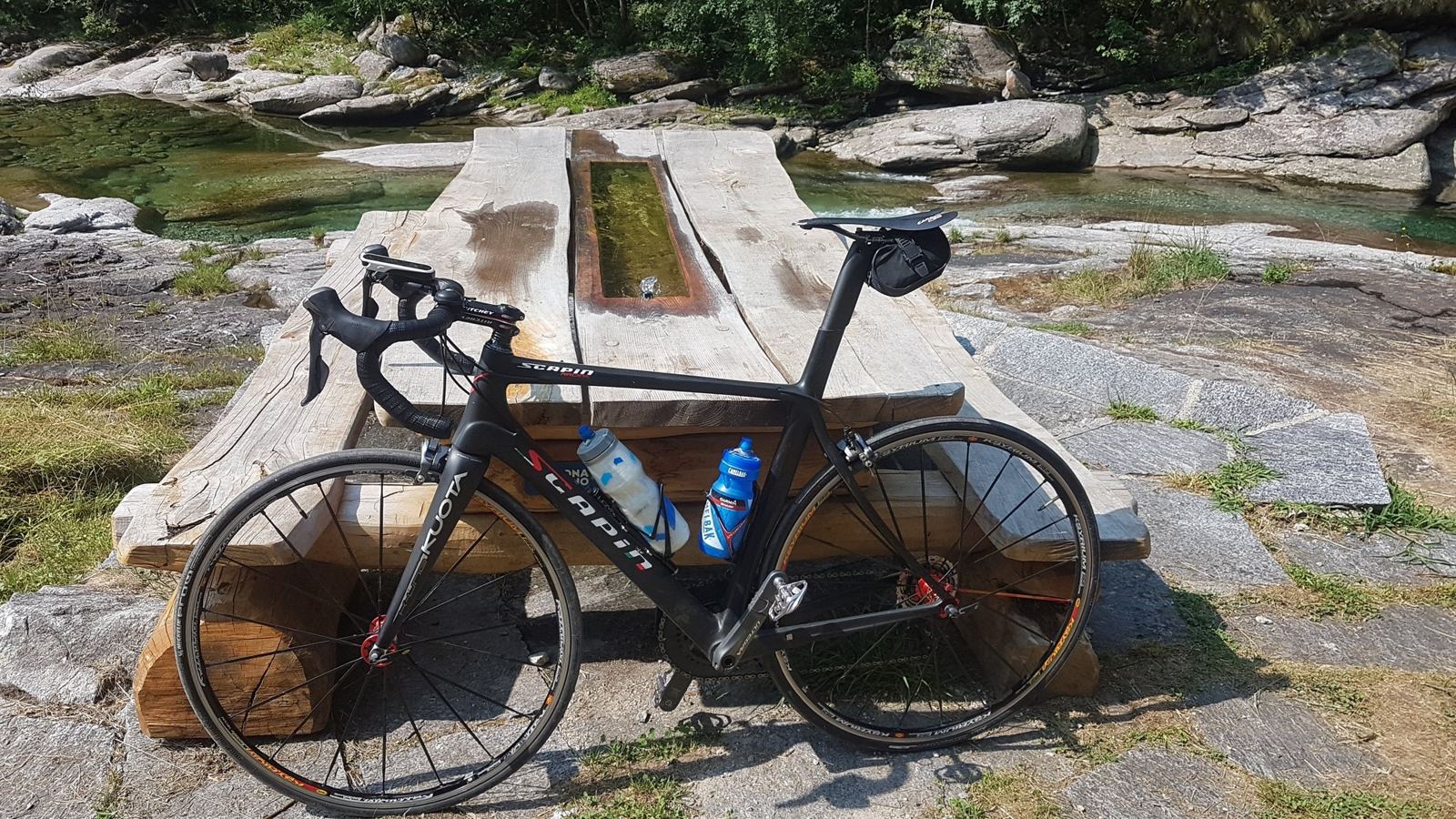 A carbon bike propped against a stone bench by a turquoise river pool