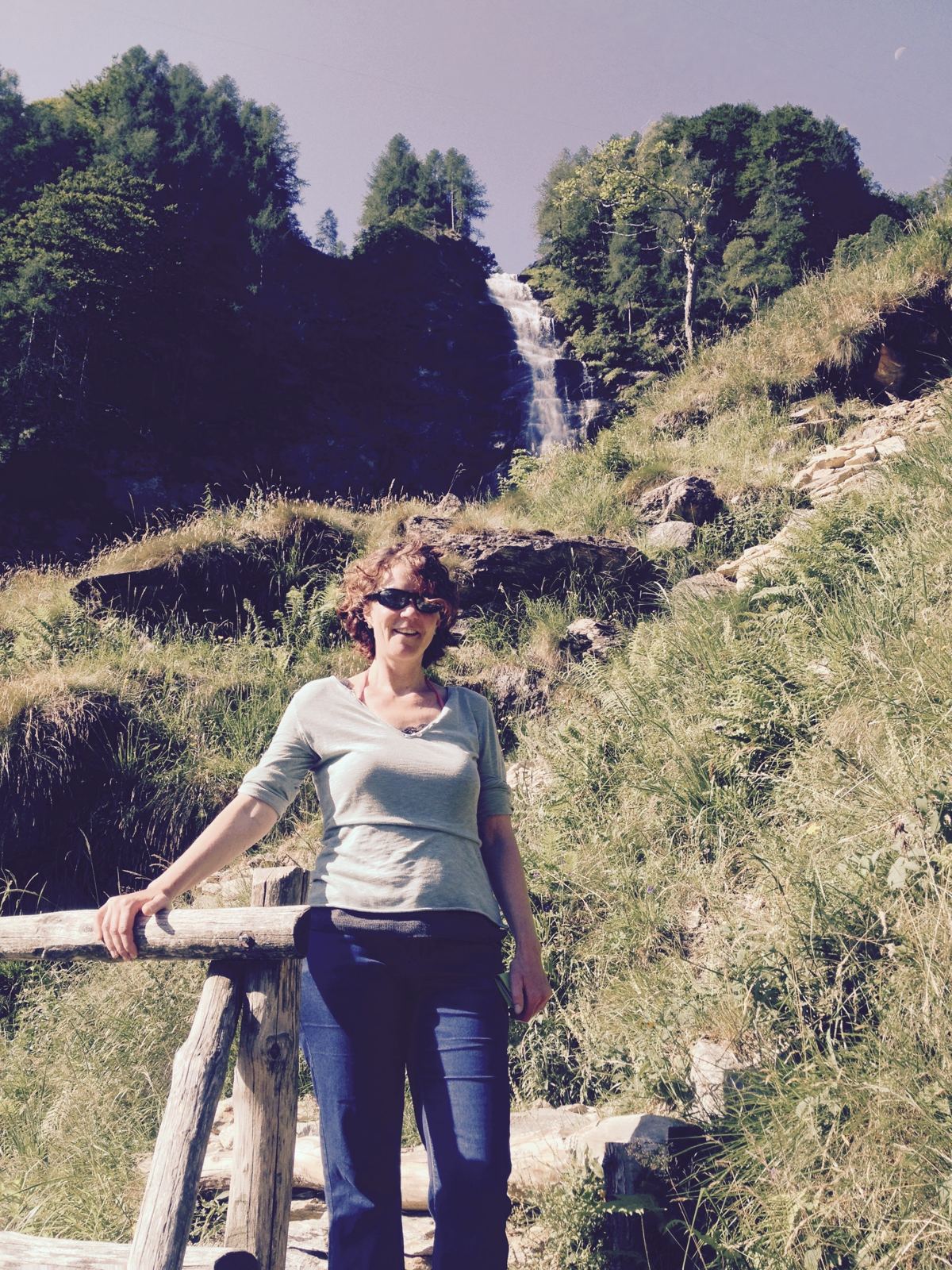 A woman standing by a wooden fence on a mountain trail near a waterfall above Sonogno