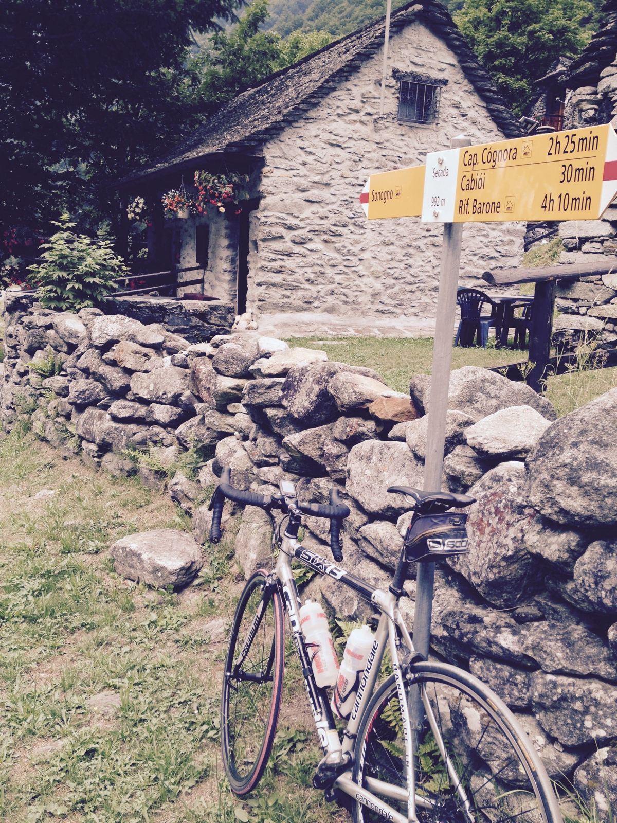 A road bike leaning against a dry stone wall in front of an old stone house with yellow hiking signposts