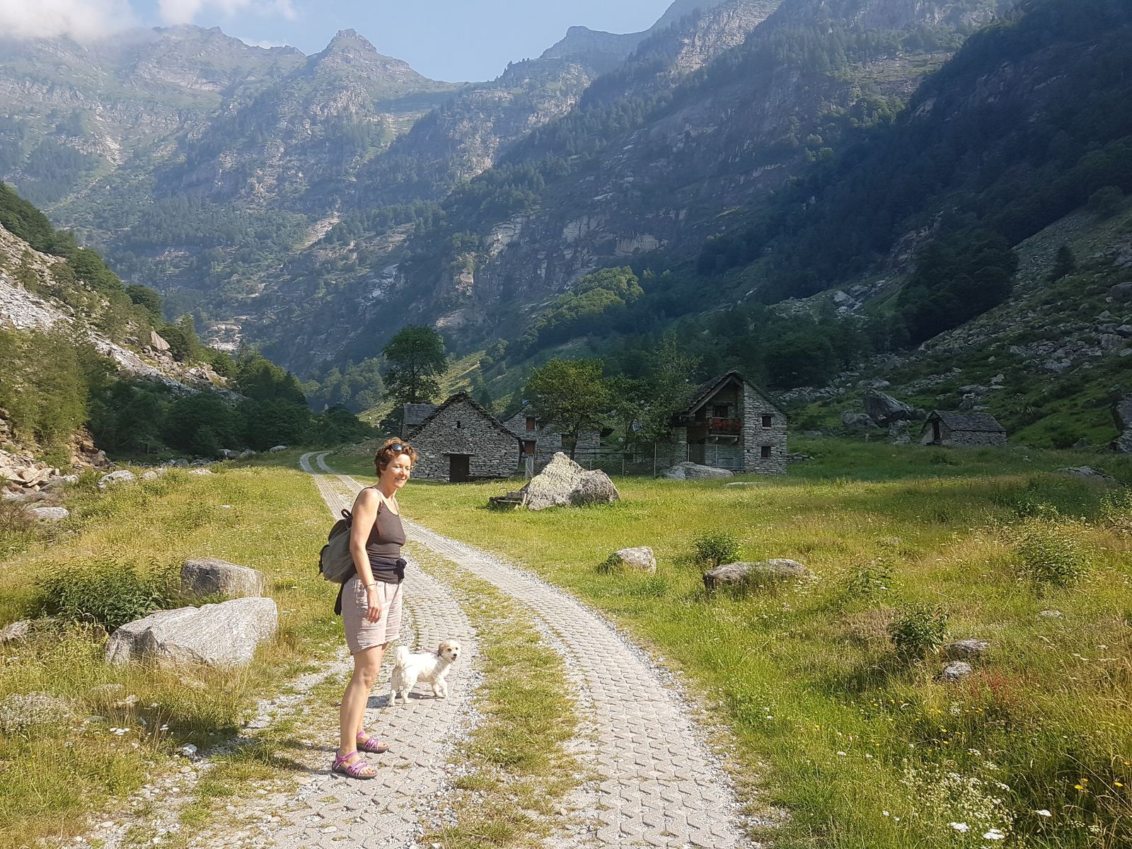 A woman and a small dog walking a gravel track towards a stone hamlet in the Verzasca valley
