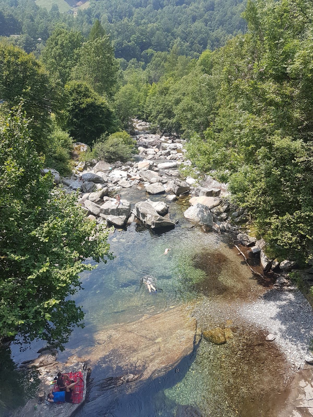 People swimming in a clear green river pool between boulders at the top of the Verzasca valley