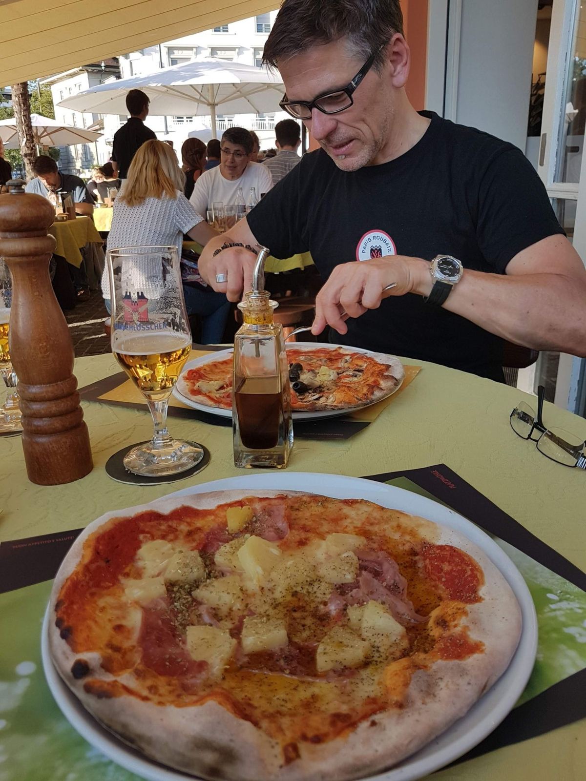 A pizza on a restaurant table with a cyclist eating at a busy terrace behind