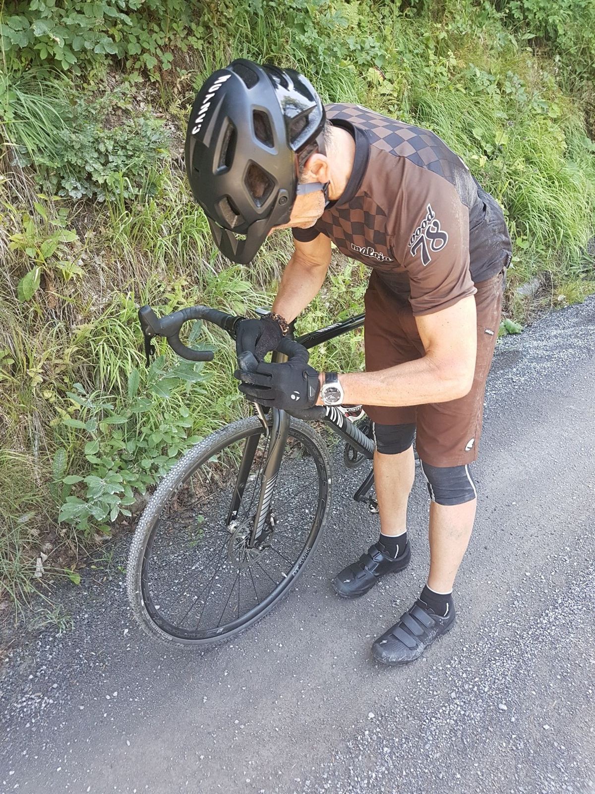 A cyclist in a helmet bending over a cross bike at the side of a gravel track beside a grassy bank